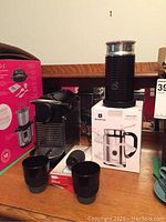 Nespresso coffee maker, Aeroccino milk frother on box, two black mugs, and Nespresso pods box on wooden surface