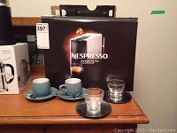 Photo of the espresso maker box, showing the Nespresso Essenza Mini model and four cups with saucers placed in front of it.
