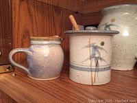 Photo showing a stoneware creamer jug and honey pot with lid and wooden dipper on wooden shelf.