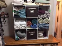Full view of white melamine 9-cube shelving filled with towels, two fabric storage bins, and cushions on a wooden table