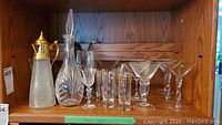 Wooden shelf displaying a tall crystal teapot-shaped decanter with gold lid and handle, clear cut-glass decanter with stopper, and an assortment of various crystal glasses including stemmed and tall glasses.