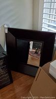 Photo showing full view of square mirror with wide dark wood frame leaning against the wall near a window and hardwood floor.