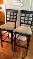 Front view of two dark wood bar stools with light beige micro suede cushions and lattice style backrests on hardwood floor near kitchen.