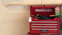 Front view of closed Mastercraft red metal tool chest with a cardboard label and some tools on top including gloves and steel wool.