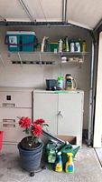 Full view of shelving with Rubbermaid ice chest, garden sprays, fertilizers, and other garden items on shelf. Below shelf is white cabinet and large ceramic pot with red artificial flowers, fertilizers and watering cans on the floor in front.