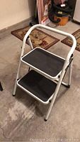 White metal two-step folding step stool with black textured steps seen standing open on concrete floor.