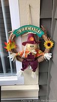 Fall-themed decorative wreath with smiling scarecrow, 'Welcome' sign, and autumn leaves, hanging wreath style.