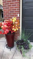 Faux maple plant with red and yellow leaves in tall red planter on metal stand, placed outdoors by brick wall and garage door.