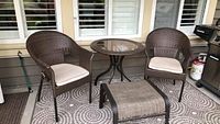 Two brown resin wicker-style outdoor armchairs with beige cushions, round glass-top table, brown padded footstool, and outdoor patterned rug underneath.