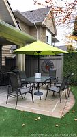 Full view of the patio set including table, six chairs, and green umbrella on a patio with artificial grass and concrete tiles.