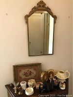 Full view of gilded wooden framed mirror above table with other items including needlepoint, cameo frames, teapot, and other decorative pieces