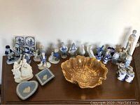 Various porcelain figurines arranged on table including Lladro swans, Blue Delft tiles, Wedgewood dishes, and other figurines with flower-shaped bowl