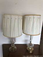 Two vintage crystal table lamps with pleated white fabric shades trimmed with gold braid sitting on a wooden surface against a white wall.