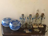Wide shot showing two candlestick style lights with hanging blue and clear plastic crystals, two blue painted glass hurricane globes with painted white landscapes, extra crystals laid out on wooden surface