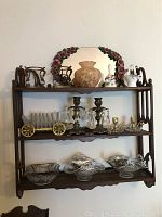 Full view of dark wood three-tier wall shelf displaying candle holders, crystal and brass coaster set, antique glass dishes, ceramic vase, floral wreath, and small pitchers