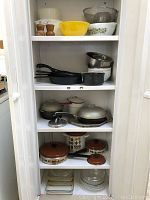 Full view of white shelving unit containing vintage kitchen items including cast iron skillets, metal mixing bowls, brown lidded pots, and glass bakeware.