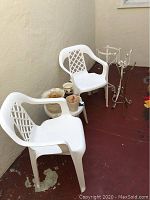 White plastic patio chairs and vintage metal plant stands with garden pots placed around them on a red floor against light wall.