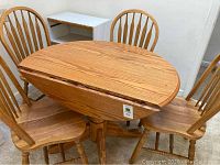Full view of oak drop leaf kitchen table with four matching chairs arranged around it, showing the wood grain and the table with one leaf folded down.