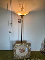 Full view of brass floor lamp with needlepoint framed artwork at base