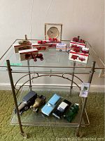 Full view of the drink cart with glass shelves and an assortment of model cars and mini trucks on both shelves.