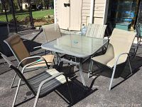 Photo showing patio square table with glass top and five surrounding patio chairs in beige mesh fabric with two chairs having striped fabric backs, all outdoors on black paved surface.