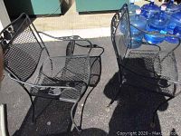 Two black wrought iron chairs with mesh seats and backs seen from a corner angle outside. Chairs have curved armrests and visible wear.