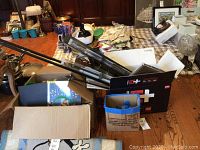 Boxes containing several bought curtain rods, a small wooden shelf and a blue box full of cabinet knobs, all items on a carpeted floor with other household items in the background.