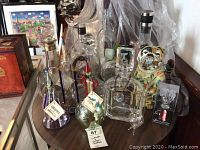 Photo showing a table with a variety of vintage and unique glass bottles, decanters, and a ceramic bulldog bottle. Some bottles have labels and decorative stoppers.