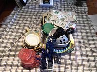 Wide view of picnic and party supplies arranged on a plaid tablecloth showing bowls, trays, kettle, containers, and linens.