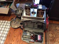 Photo showing stacked cookware including chafing dish, coffee pots, cooking pans, grilling utensils in plastic case, and boxed espresso pots.