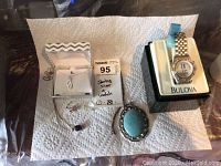 Photo of all items on white paper towel showing three necklaces, ring, turquoise pendant, and Bulova watch on display stand.