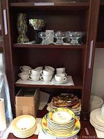 Shelves with white ceramic cups and saucers, colorful fruit design dinner plates, and clear glass footed dessert glasses.