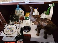 Shelf with multiple figurines including a large brown elephant statue with lifted trunk, a white ceramic elephant, a Wedgwood dish, Manufacture De Monaco trinket dish, monkey figurines and a blue round trinket box