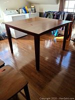 Square high dining table with a medium brown wood finish placed on hardwood floor, shows table overall structure and design.