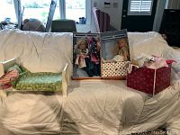 Photo showing small wooden doll loveseat and three boxes containing dolls and clothing/accessories on a couch.