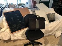Photo showing office chair with cloth seat and back, framed chalkboard, corkboard, and laptop case in front of a couch.