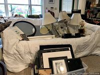 Photo showing the couch covered in white cloth with various picture frames leaning on it and stacked in front, three decorative table lamps, and small decorative items placed on the couch.