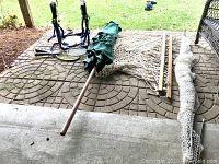 Photo showing green patio umbrella with wooden pole, foldable bike rack, natural rope hammock with wooden spreader bars, rolled golf practice net, and tennis rackets all placed on patio mat outside.