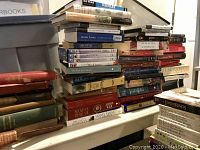 Wide view of mixed collection of stacked books and DVDs on a white table with a grey storage bin in background.