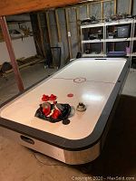 Photo showing a full-size air hockey table with white playing surface, red markings, multiple red paddles, pucks, and a black bag containing accessories placed on the table.