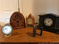Four antique clocks and a heavy iron object on wooden surface: Seth Thomas, Ingraham, black mantel clock, mid-century alarm clock, and radio replica.