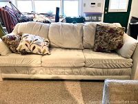 Front view of beige microfiber couch with three seat cushions and multiple decorative pillows. Couch positioned in living room with carpet underneath.