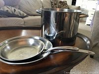 Photo showing two stainless steel skillets stacked inside each other and a large stainless steel stockpot without lid on a wooden tray.