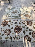 Photo showing two wool area rugs with distinct floral patterns laid flat on a wooden deck surface.