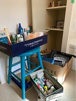 Blue stool holds a cardboard box with assorted bathroom and skincare bottles, another box on floor contains various tubes and bottles. In background, a cardboard box filled with assorted cosmetic bags.