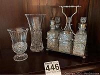 Front view of crystal cruet set with three cut glass bottles in metal carry-all alongside two small crystal vases on wood surface.