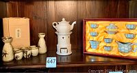 Photo shows 6 cup two server Japanese sake set with original box, white tea pot warmer, and small rectangular porcelain cup all displayed on wood shelf.