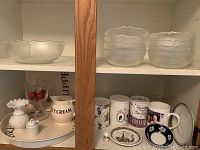 Glass serving salad bowl and eight matching smaller bowls on shelf with various mugs, a small decorative white ceramic vase, cream jug, and goblet on a serving tray.