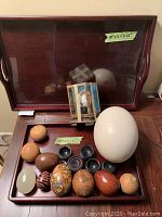 Wide shot of wooden trays filled with various wooden, stone, and painted decorative eggs, small black egg cups, and a small reverse-painted glass box. Shows size labels of trays.