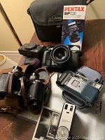 Photo showing Pentax SF10 SLR camera, leather case, leather binoculars, blue Asahi camera with case, black carrying case, and Mecablitz flash manual.